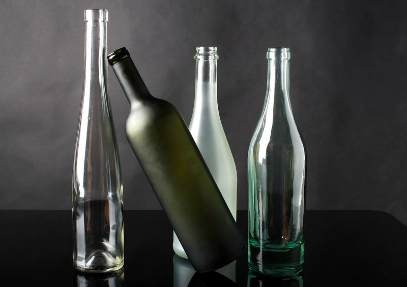 Schwarzer Bartresen vor grauem Hintergrund mit edlen Flaschen aus farblos-transparentem Glas, grün-milchigem und weiß-milchigem Glas darauf und deren Spiegelung in der Tresenfläche
